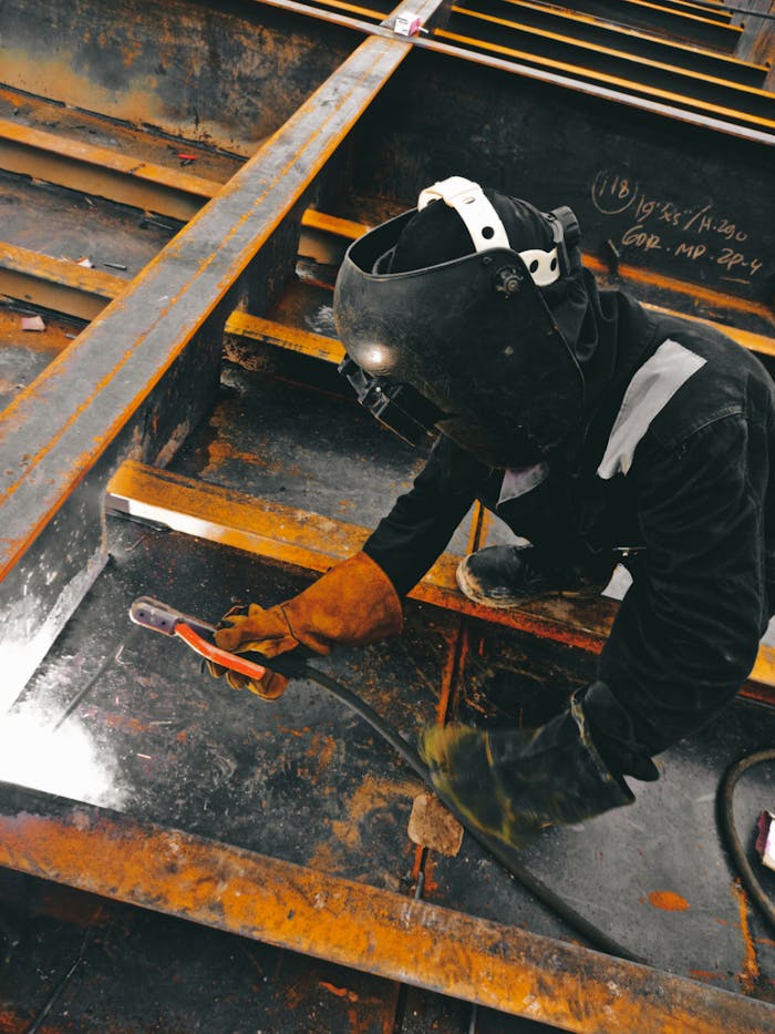 gallery-5 A welder in action wearing protective gear and welding helmet in an industrial setting.