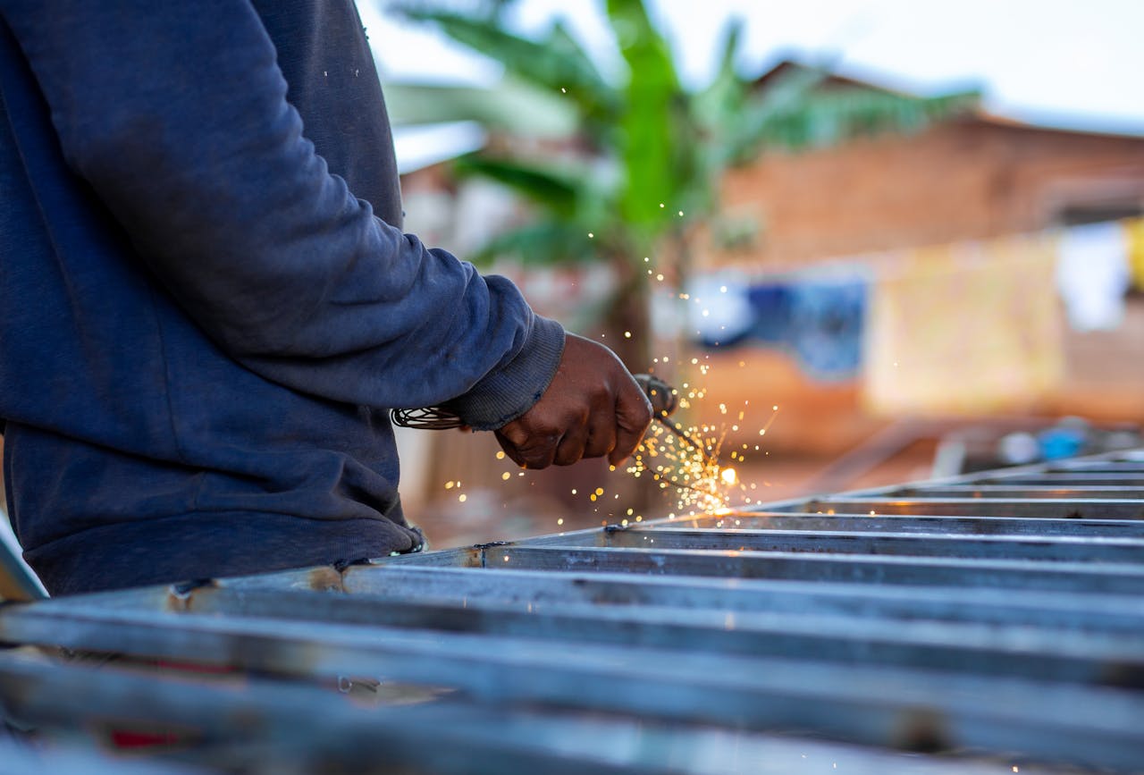 gallery-4 A person welding metal outdoors, creating bright sparks against a blurred background.