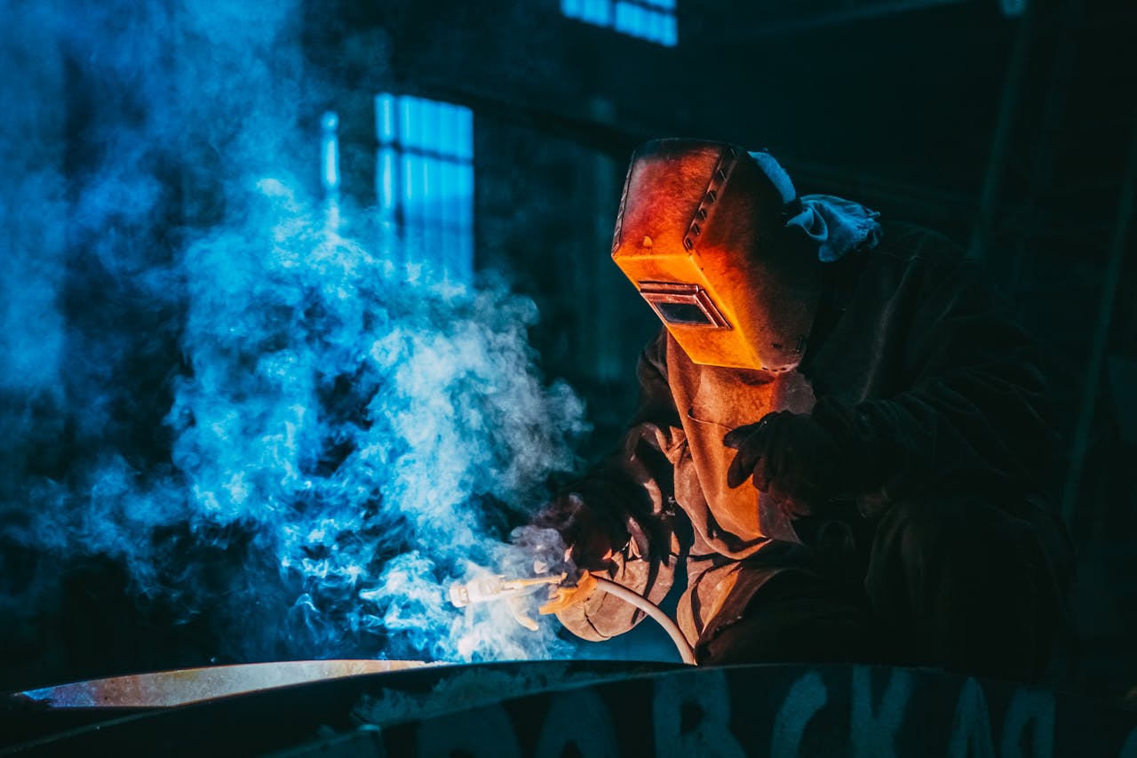 services-01 A welder in a workshop using safety gear to weld metal, surrounded by blue smoke and atmospheric lighting.